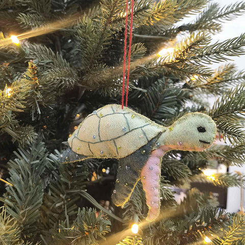 A handmade felt sea turtle ornament with a light green shell, blue-gray flippers, a pale pink underside, and a red hanging string, shown against a plain white background.