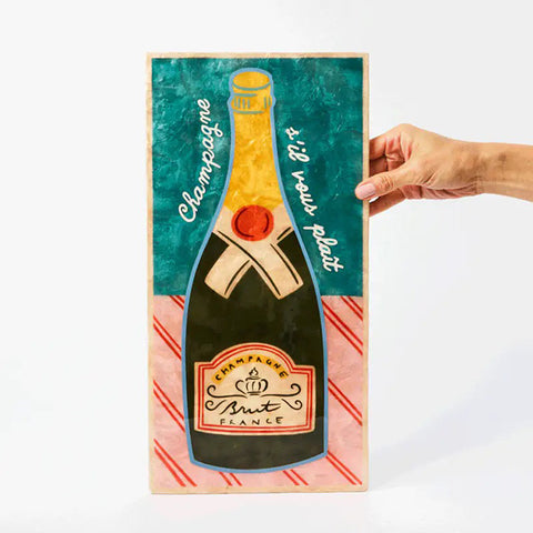 Decorative sign with a champagne bottle design next to a cocktail and red napkin on a white surface.