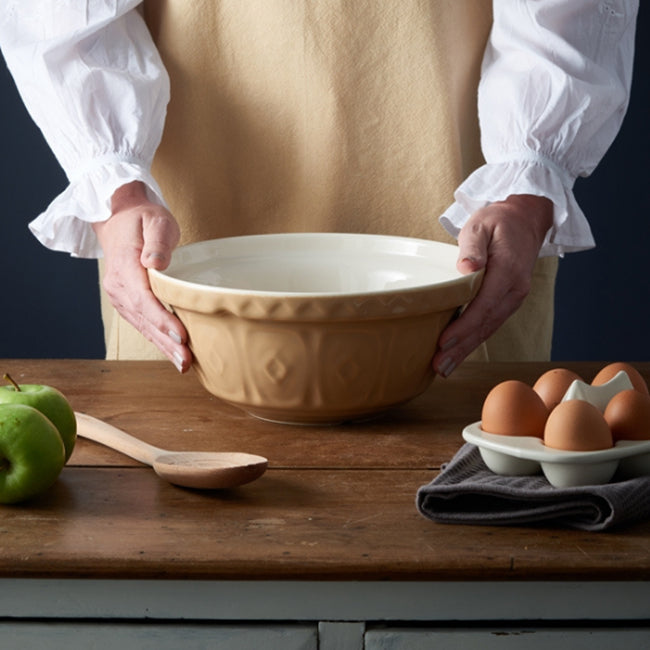 Mason Cash Cane Mixing Bowl on bench with eggs wooden spoon and apples, a person is holding the bowl wearing an apron 