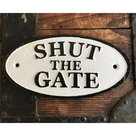 An oval white metal sign with black lettering that reads 'SHUT THE GATE', mounted on a rustic dark brown wooden surface.