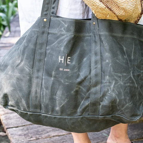 Green tote bag with 'HE' branding on a wooden hook against a concrete wall.