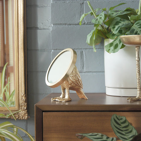 Decorative mirror with bird body on a stack of books with a plant and glass in the background